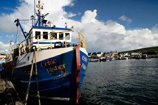 Blau-weißes Fischerboot mit dem Namen Celtic Fisher im Hafen, im Hintergrund ein Ort am Wasser