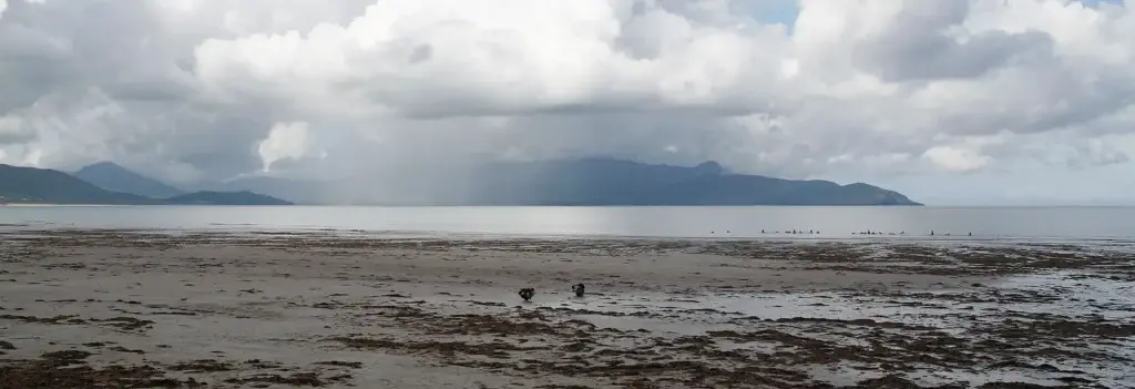 Weiter Strand bei Ebbe, in der Ferne Berge und ein Regenschauer über dem Meer