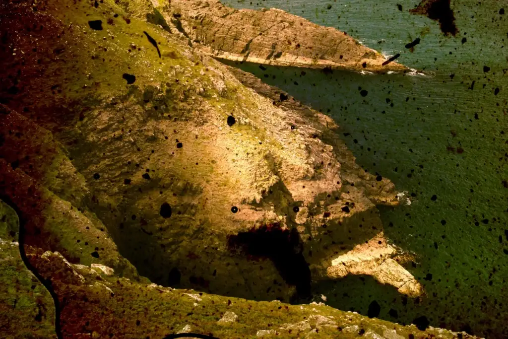 Felsige Landzunge über grünem Wasser in goldenem Licht, dunkle Flecken im Bild